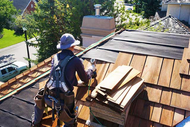 A photo of a worker installing cedar shingles on a roof for a roofing job. The worker is wearing safety gear such as a harness, helmet and gloves, and is using a nail gun to fasten the wood shingles to the roof structure.