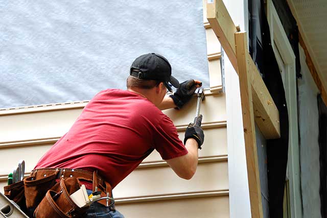A photo of a worker installing new vinyl siding on a house. The worker is holding a long piece of vinyl siding against the side of the house. The siding is a light colour and has a smooth texture.