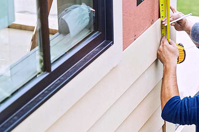 A photo of a worker using a measuring tape to measure the length of wood siding before installation. The worker is holding the measuring tape against a piece of wood and is using a pencil to mark where the wood needs to be cut.