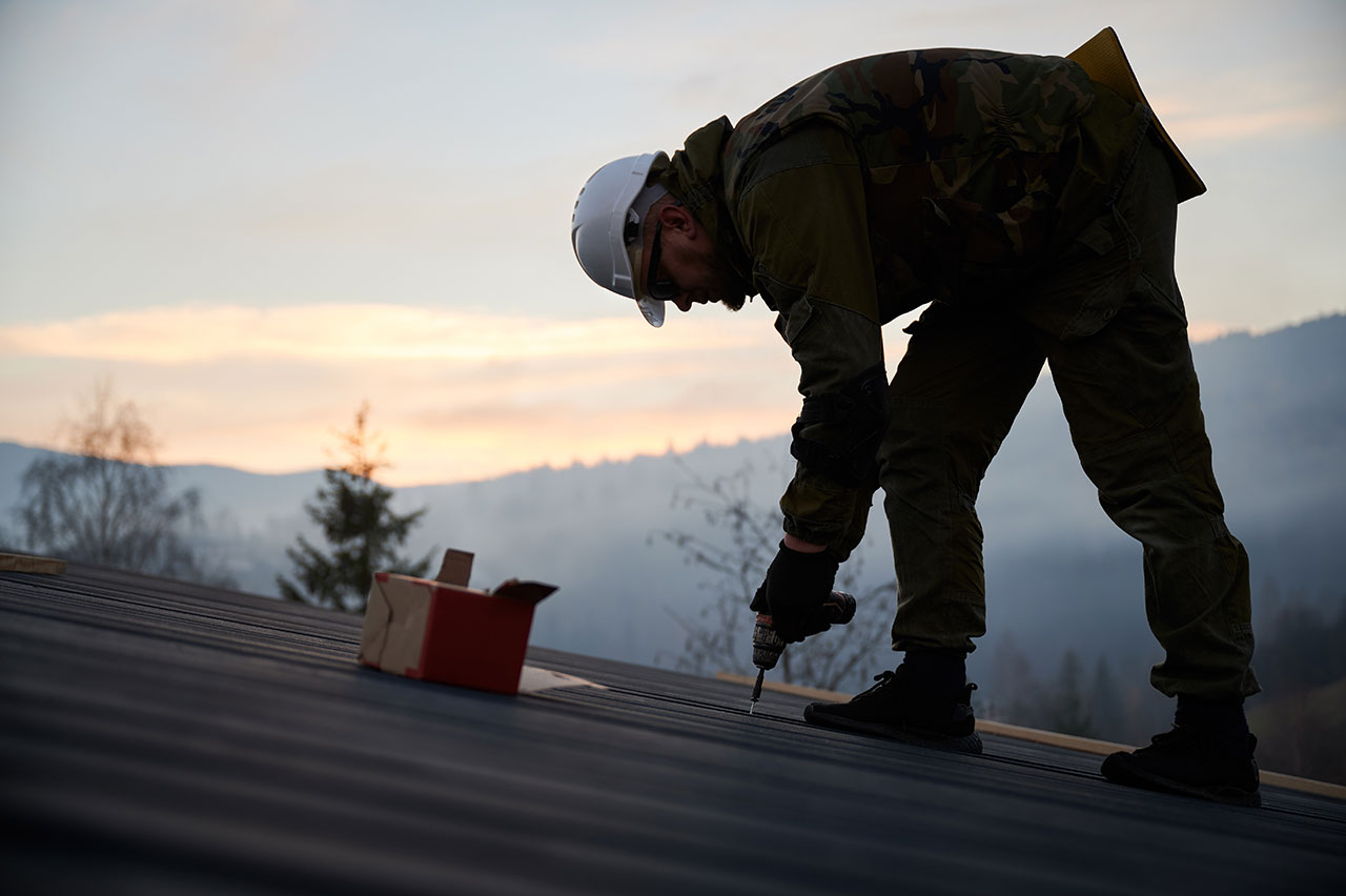 A photo of a roofer using a drill to install plywood on a roof during a Calgary roof repair. The roofer is standing on the sloping roof and is holding a drill with two hands. The plywood is being screwed into the roof's structure as part of the repair process.