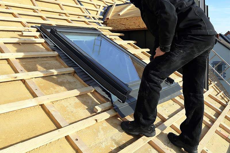 An image of a roofer inspecting a leaky skylight during a Calgary roof repair. The roofer is standing on the roof and is leaning over a skylight to examine it closely. The skylight is surrounded by flashing, and the roofer is checking for any signs of damage or wear that could be causing the leak.