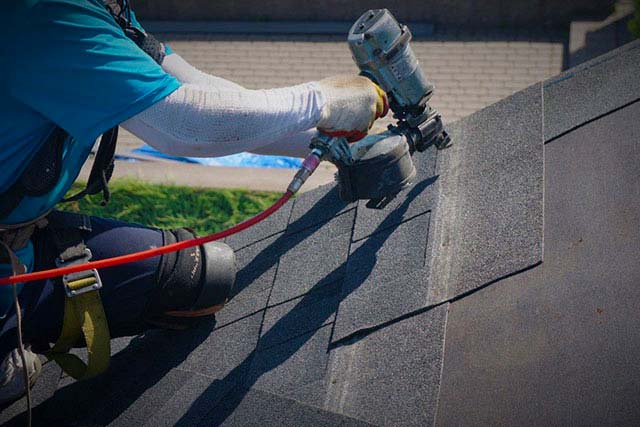 A photo of a roofer in the midst of a roof replacement, using a nail gun to install asphalt shingles.